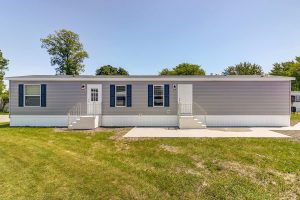 Gray single-story manufactured home with white trim, two entrances, and small steps, on a grassy lawn.