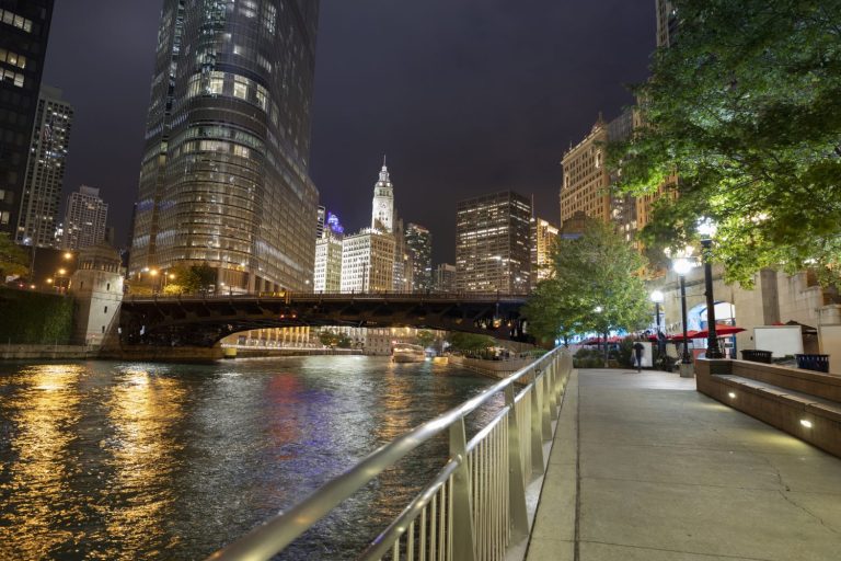 Night view of a city riverwalk, tall buildings, and a lit bridge reflecting on the water.