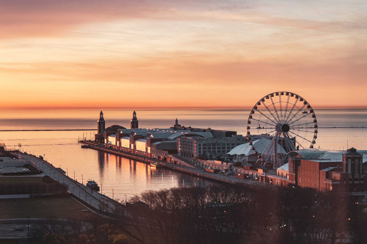 Navy Pier at sunrise with a Ferris wheel and calm lake waters, orange and pink sky in the background.