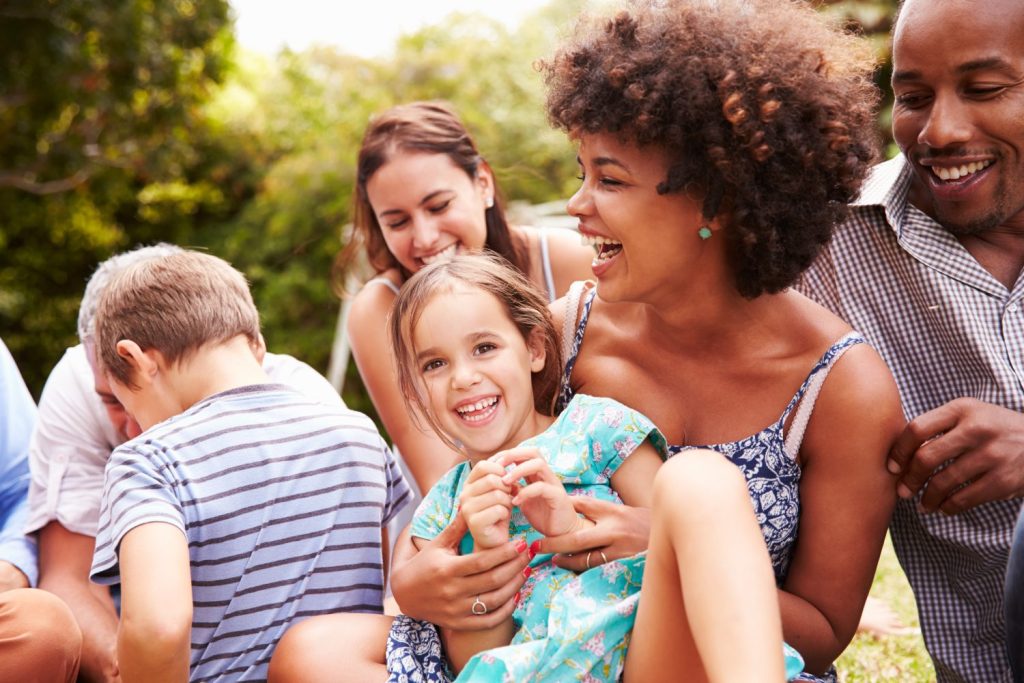 Adults and children sitting together outdoors, laughing and enjoying a sunny day in a park or garden.