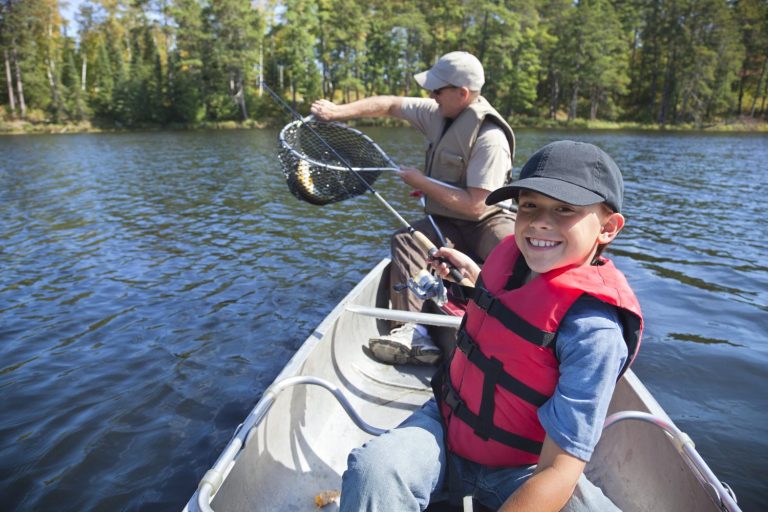 Smiling boy in a red life jacket fishing in a canoe with an adult on a lake surrounded by trees.