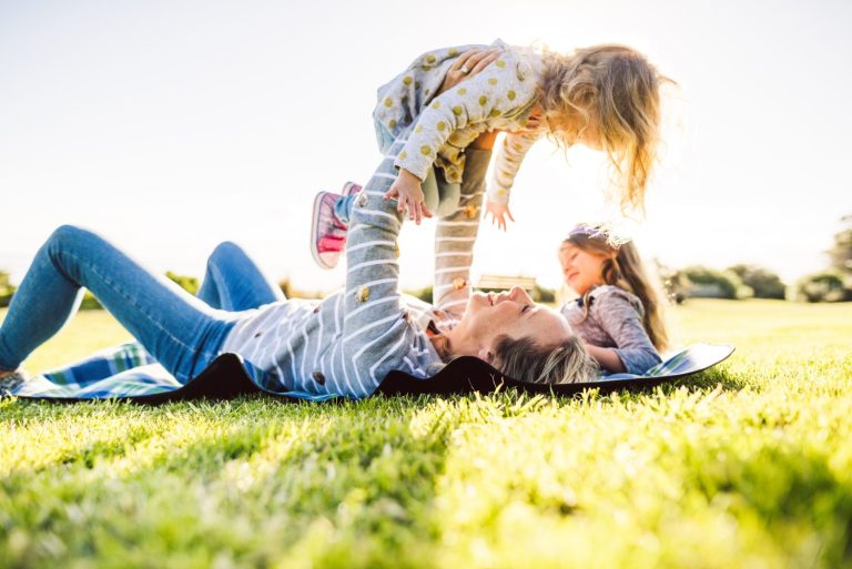 Adult lying on grass, lifting a smiling child while another child sits nearby on a sunny day in the park.