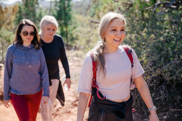 Three women hiking on a sunny trail, smiling and surrounded by trees and greenery.