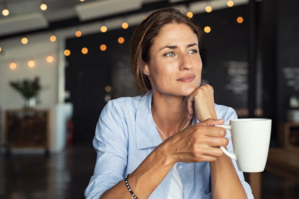 Woman in a light blue shirt holding a white mug, looking thoughtful in a cozy cafe with string lights.