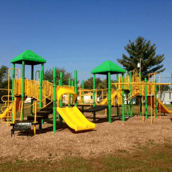 Colorful playground with green and yellow slides, bridges, and climbing areas on wood chips under a clear blue sky.