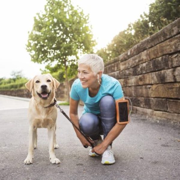 Smiling woman with short gray hair kneeling beside a Labrador retriever on a sunny sidewalk, both looking happy.